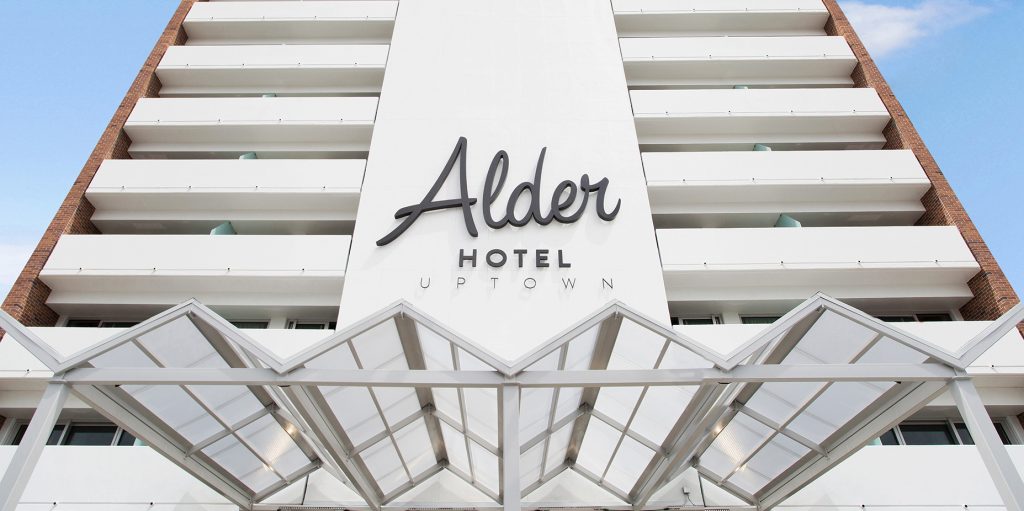 A white hotel building with balconies and a glass roof, showing the hotel name 'Alder Hotel Uptown' on its facade.